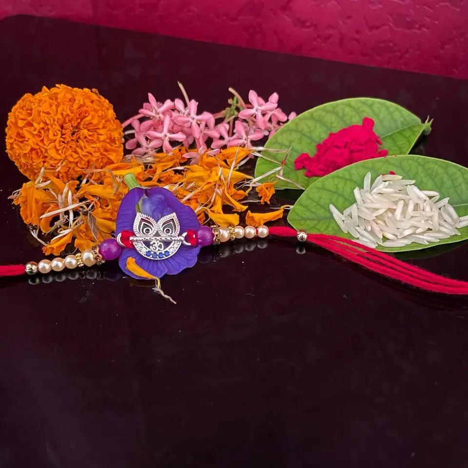 A traditional Indian Rakhi with a purple owl charm, decorated with orange marigold, pink flowers, green leaves containing rice and red powder, and a red tassel, displayed on a dark reflective surface against a textured magenta background.