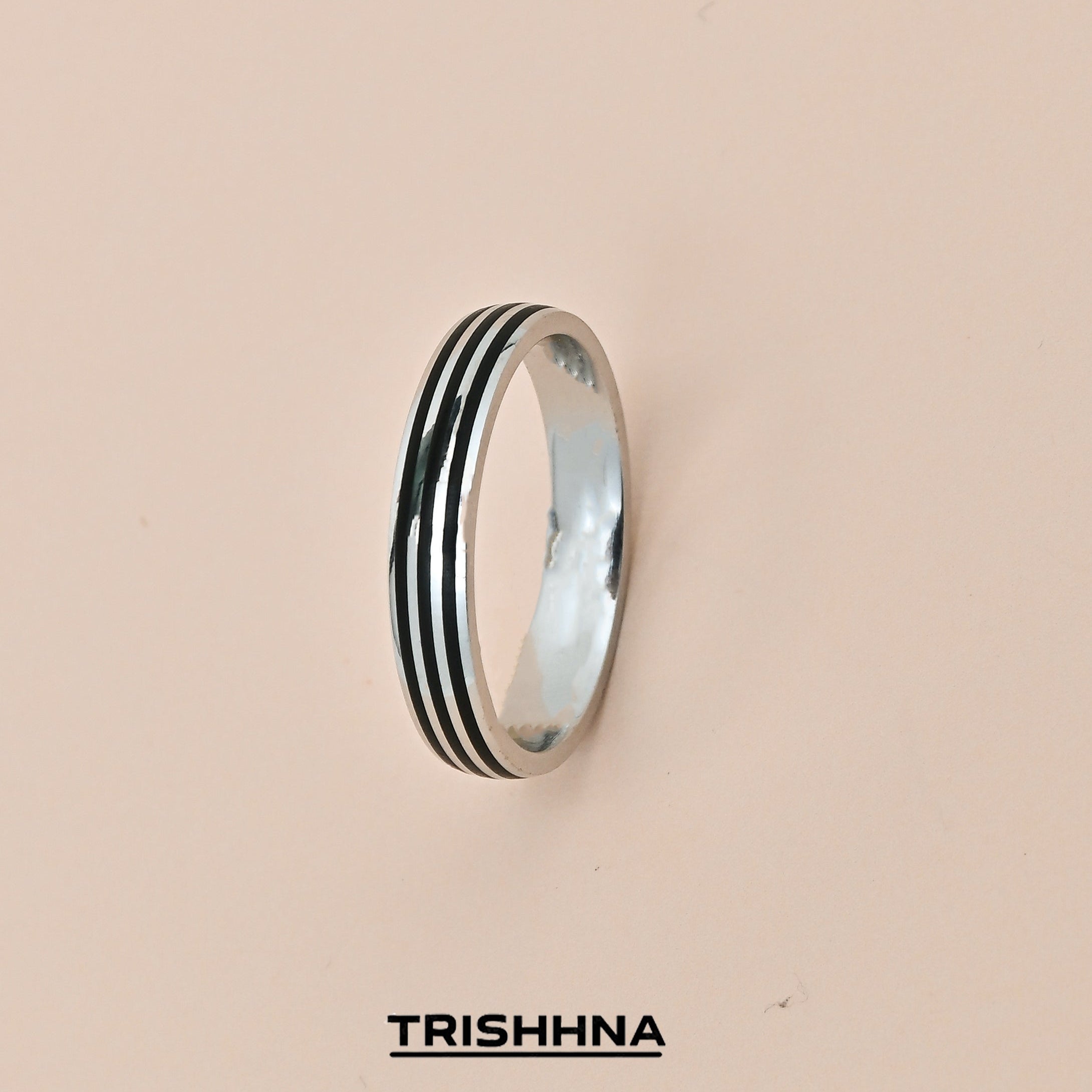 Silver ring with black stripes on a beige background, branded 'TRISHHNA'.