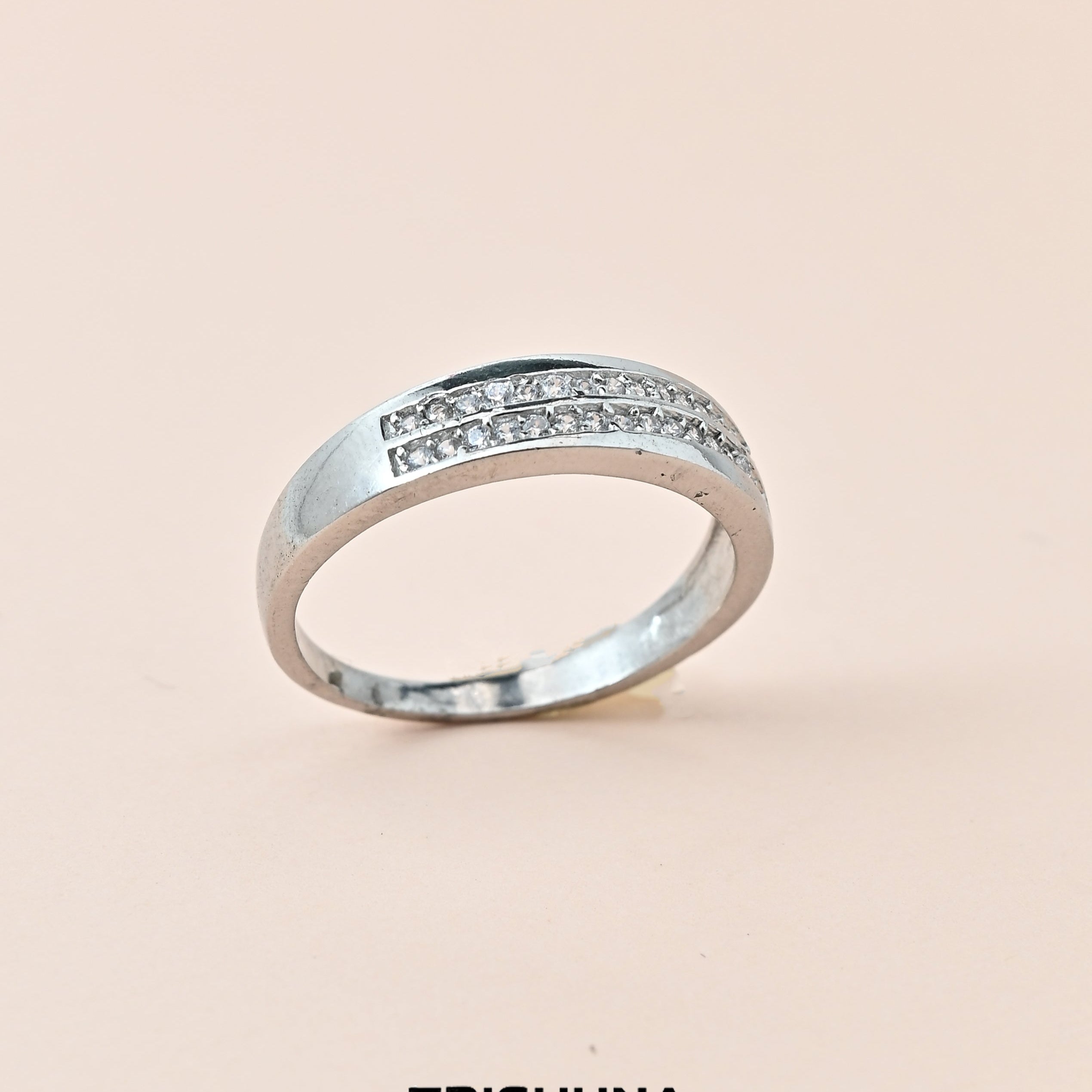 Silver ring with embedded stones on a beige background, branded 'TRISHNNA'.