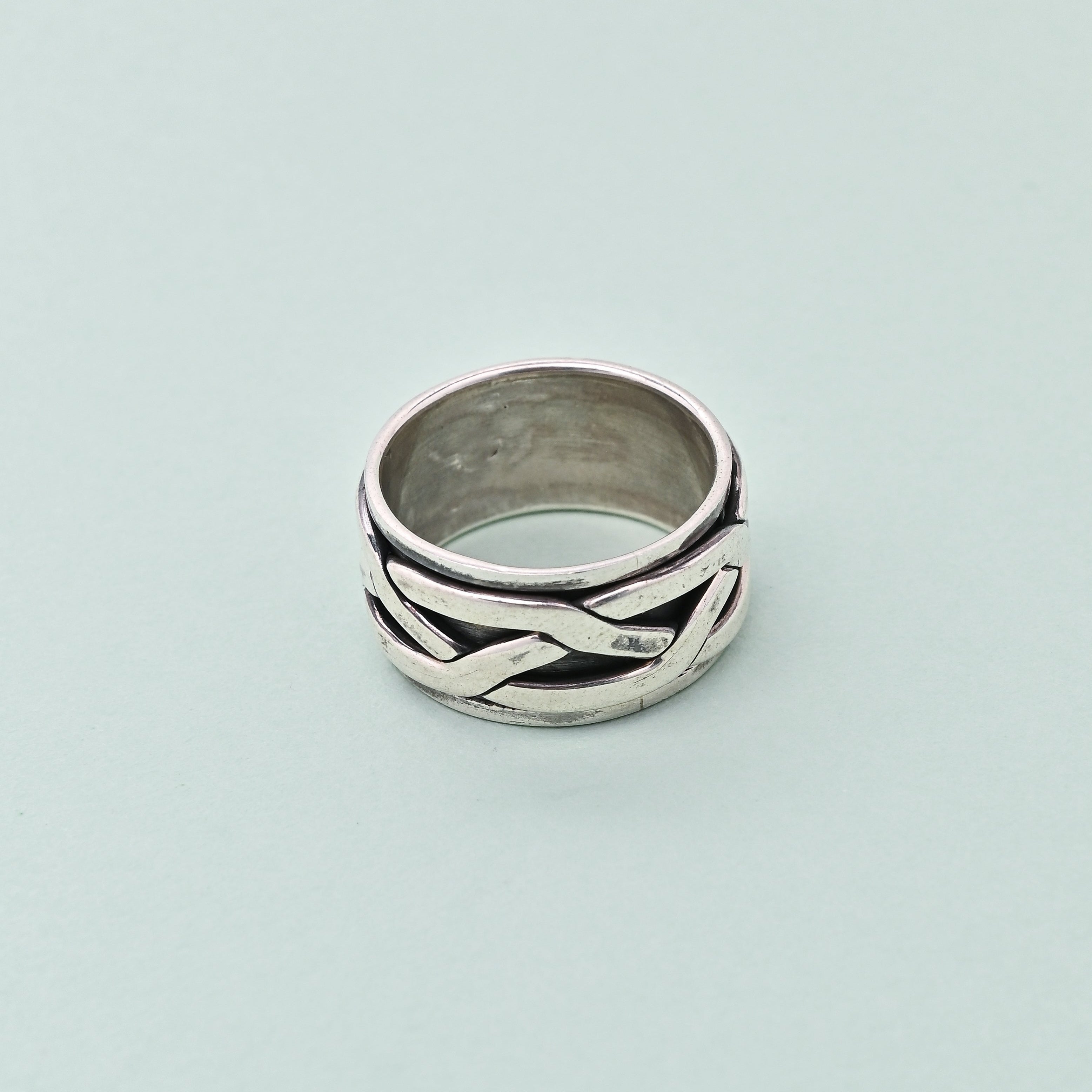 Silver ring with braided design on a light blue background, branded 'TRISHNHA'.