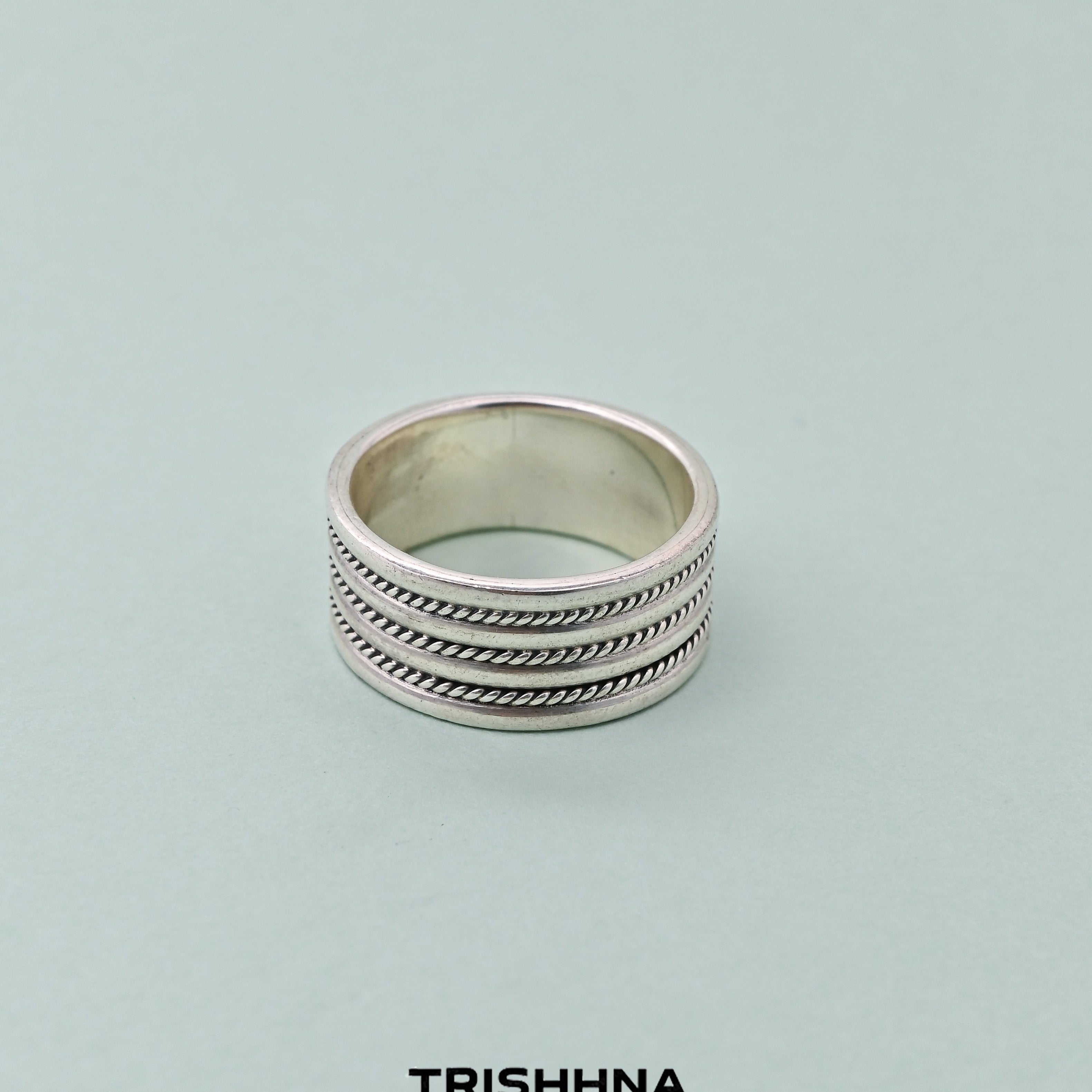 Silver ring with textured design on a light green background, branded 'TRISHNHA'.