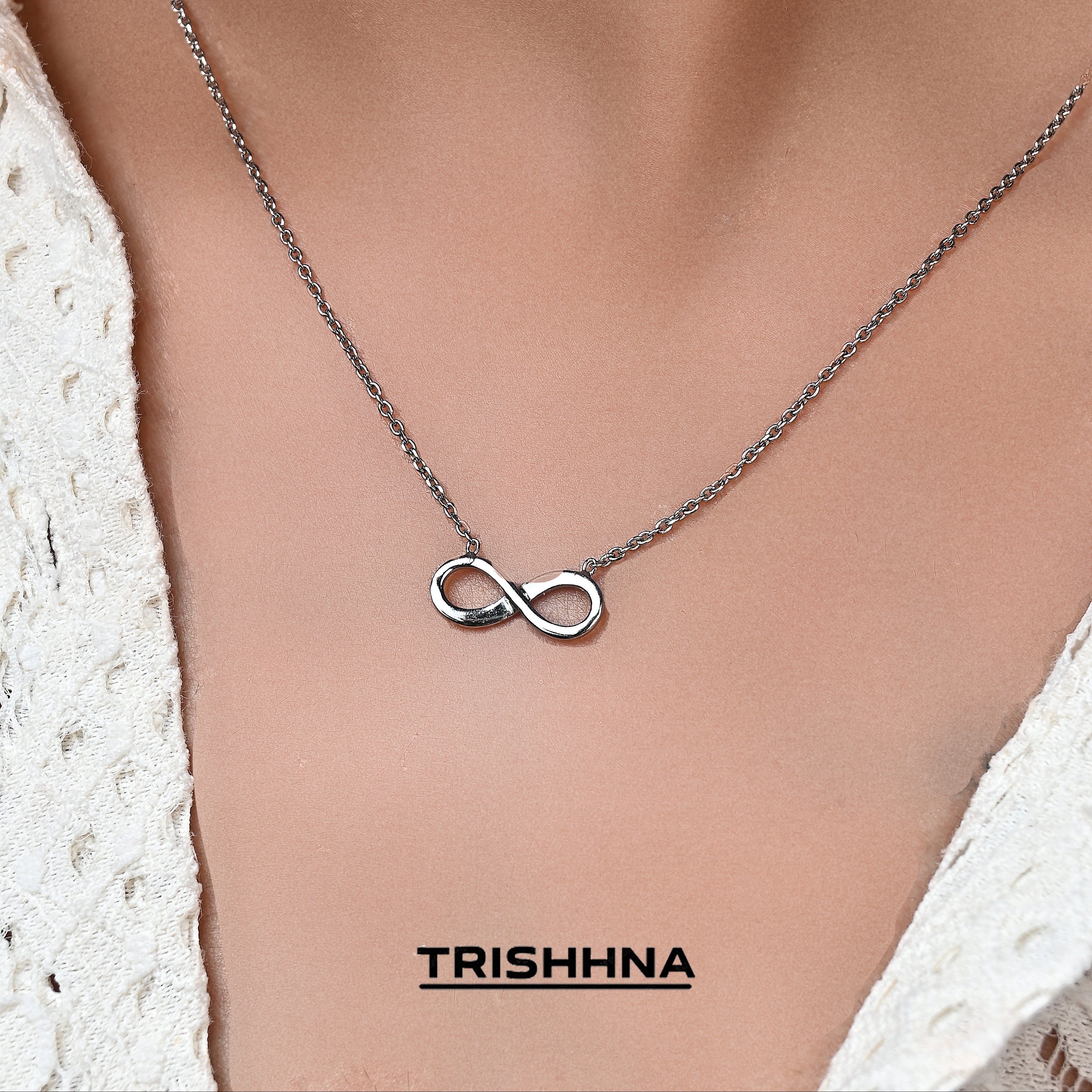 Necklace with an infinity symbol pendant on a person wearing a white lace top, branded 'TRISHNNA'.