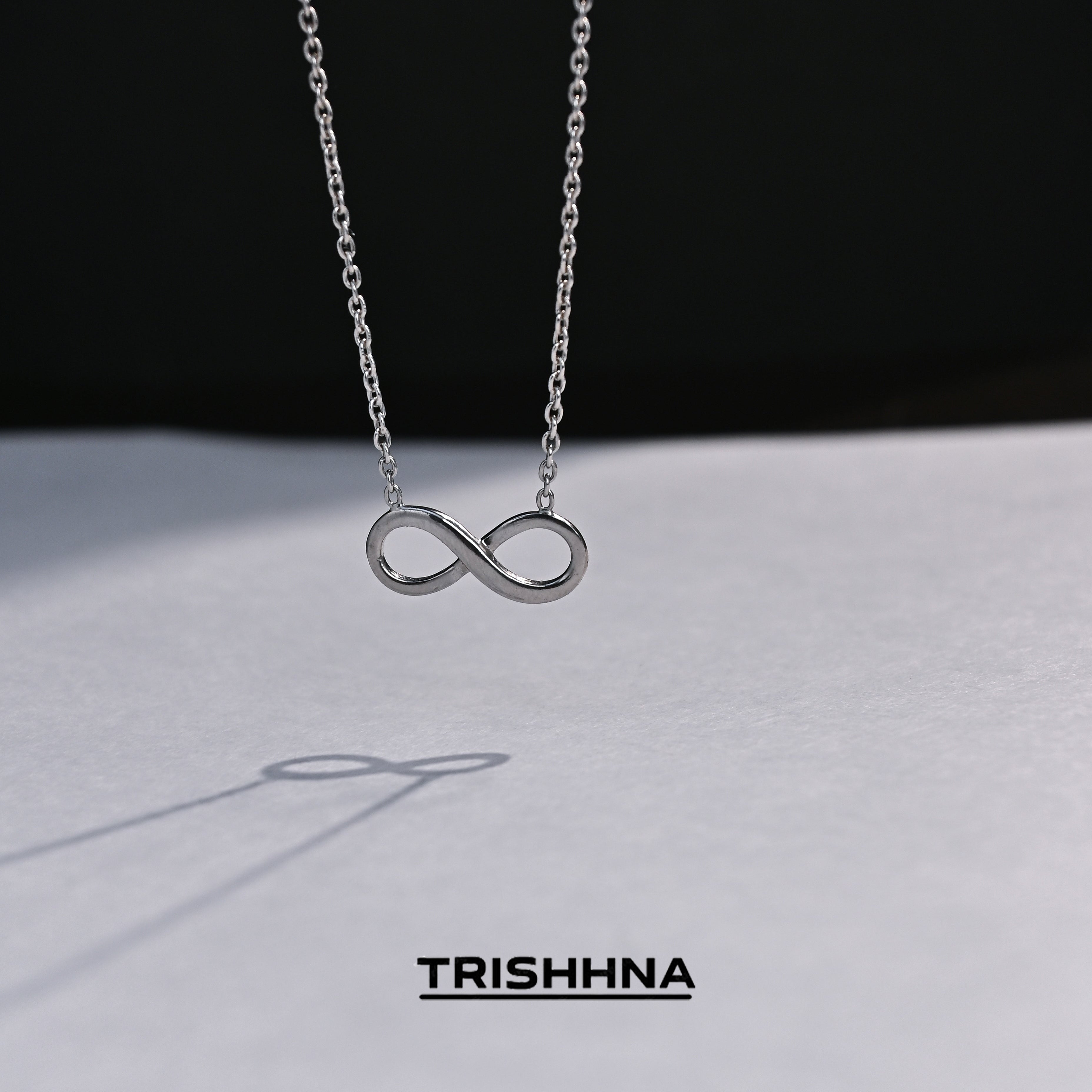 Silver infinity necklace on a dark background with 'TRISHNHA' branding.