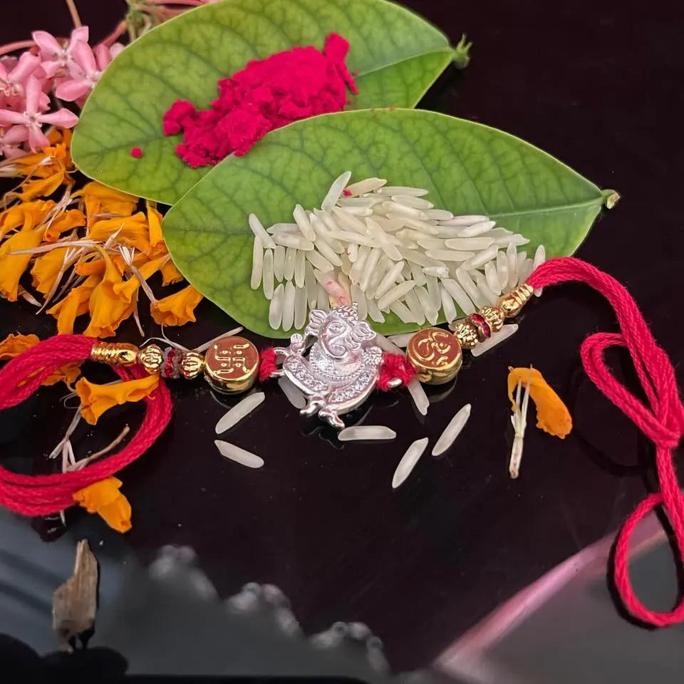 A handmade silver-toned Lakshmi pendant rakhi with red and gold thread details, displayed on two green leaves with white rice and pink and orange flower petals on a dark reflective surface.
