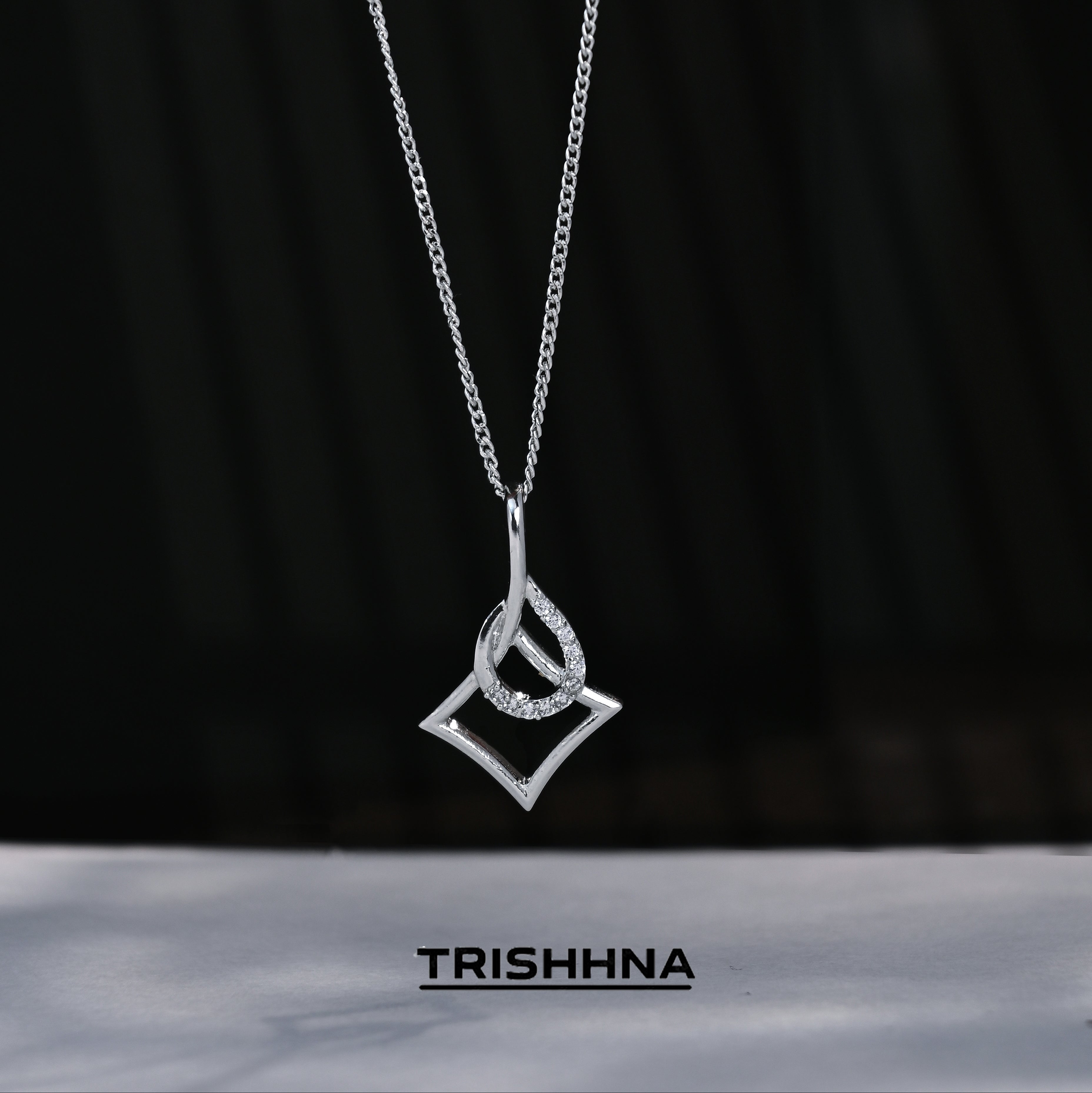 Silver necklace with a geometric pendant on a black background, featuring the brand 'TRISHNHA'.