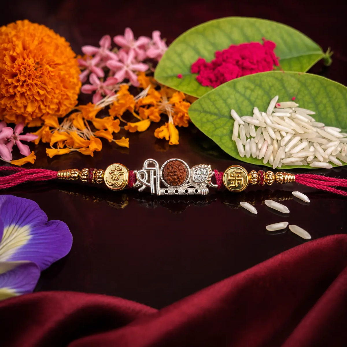 A traditional Indian Rakhi with a silver and gold-toned central charm, displayed on a dark reflective surface surrounded by marigold flowers, rice, red powder, and purple flower petals.