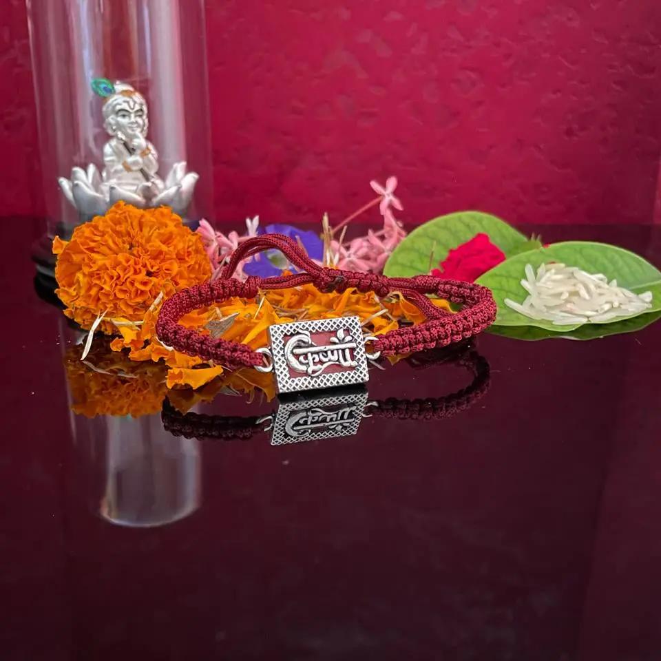 A red braided cord bracelet with a silver-toned rectangular charm engraved with Devanagari script, displayed on a dark reflective surface with orange marigold flowers, green leaves, and a small silver figurine under a glass dome against a deep red background.