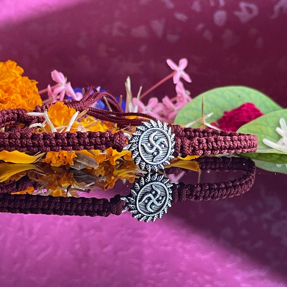 A close-up of a dark red braided cord bracelet with a silver-toned circular charm featuring a swastika symbol, styled with dried orange and pink flowers on a textured magenta background.