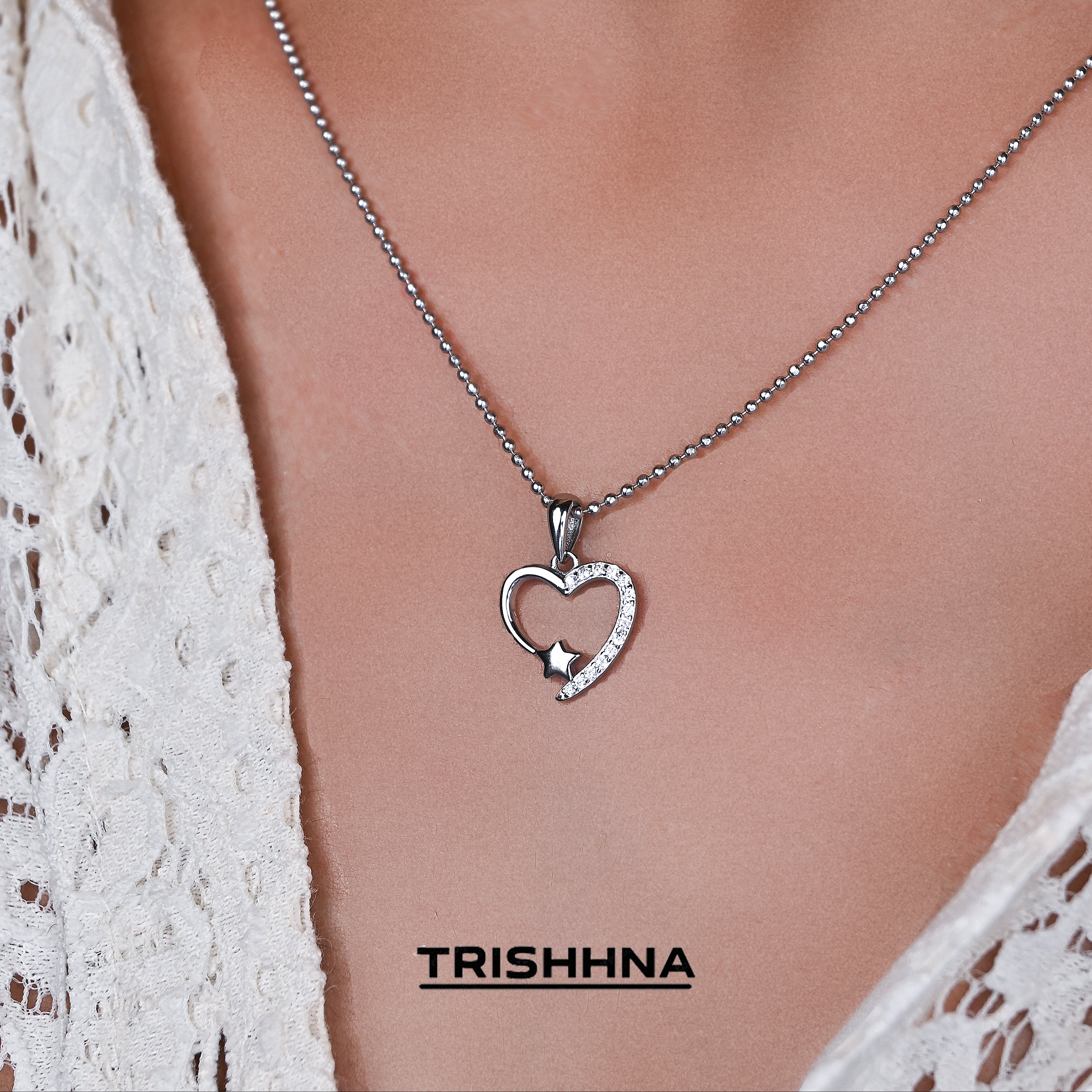 Heart-shaped pendant necklace on a person wearing a white lace garment, with 'TRISHNNA' branding.