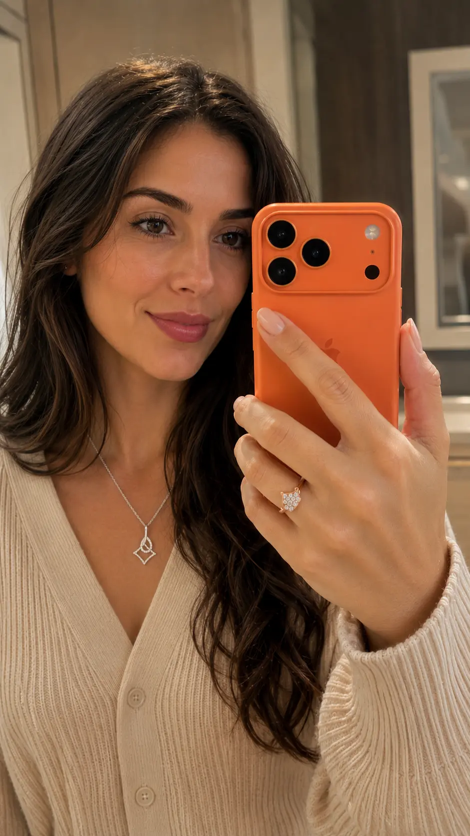 Woman holding an orange phone in a mirror selfie, wearing a beige cardigan.