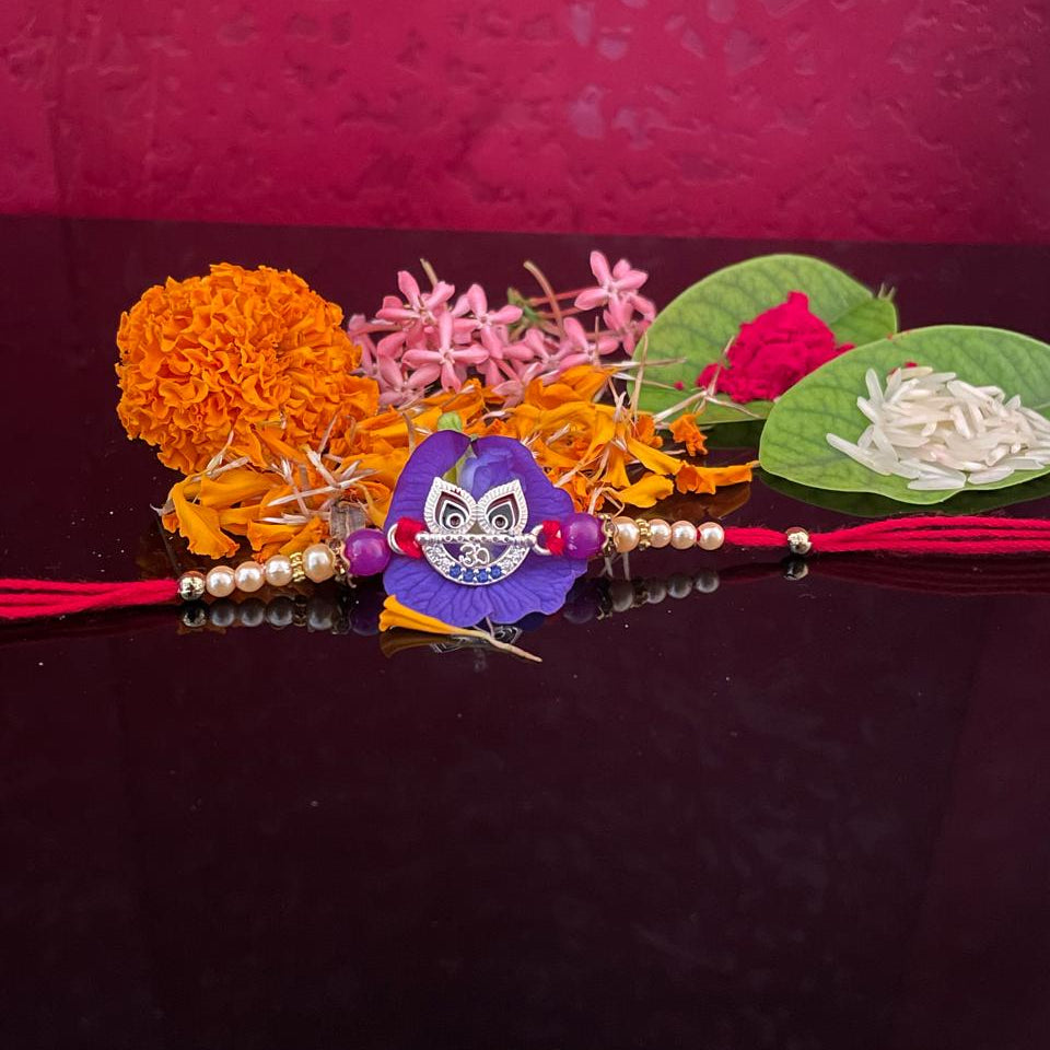 A traditional Indian Rakhi with a purple paper doll charm, decorated with orange marigold flowers, pink blossoms, green leaves, and red thread, displayed on a dark reflective surface against a textured magenta background.