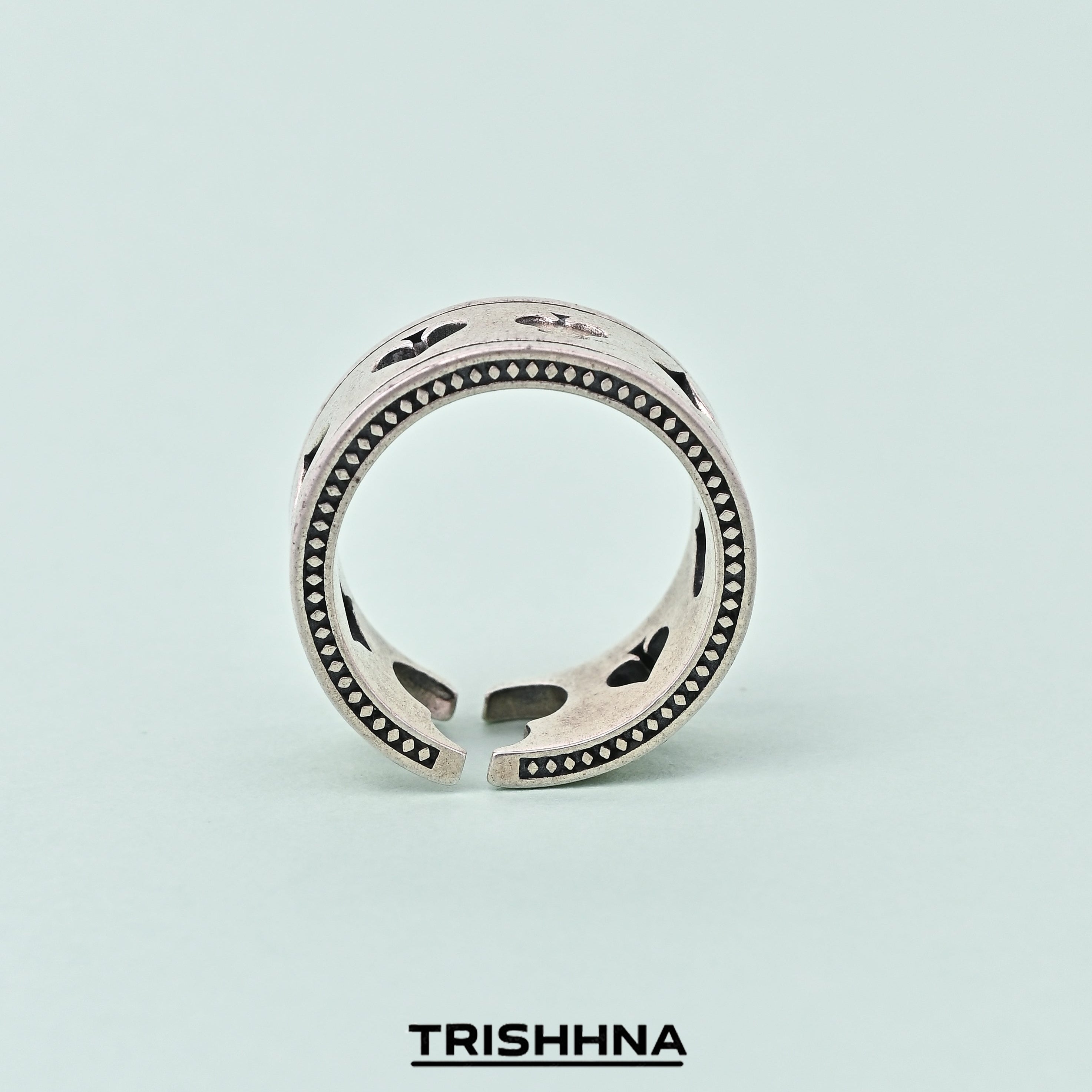 Silver ring with black accents on a light blue background, branded 'TRISHNNA'.