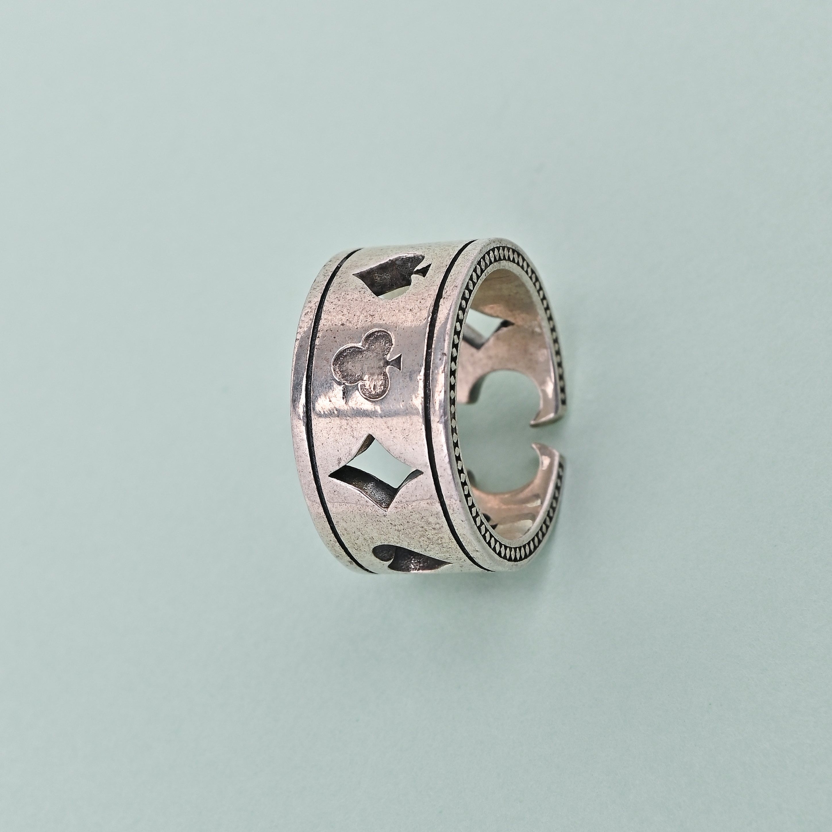Silver ring with playing card suits on a light blue background, branded 'TRISHNHA'.