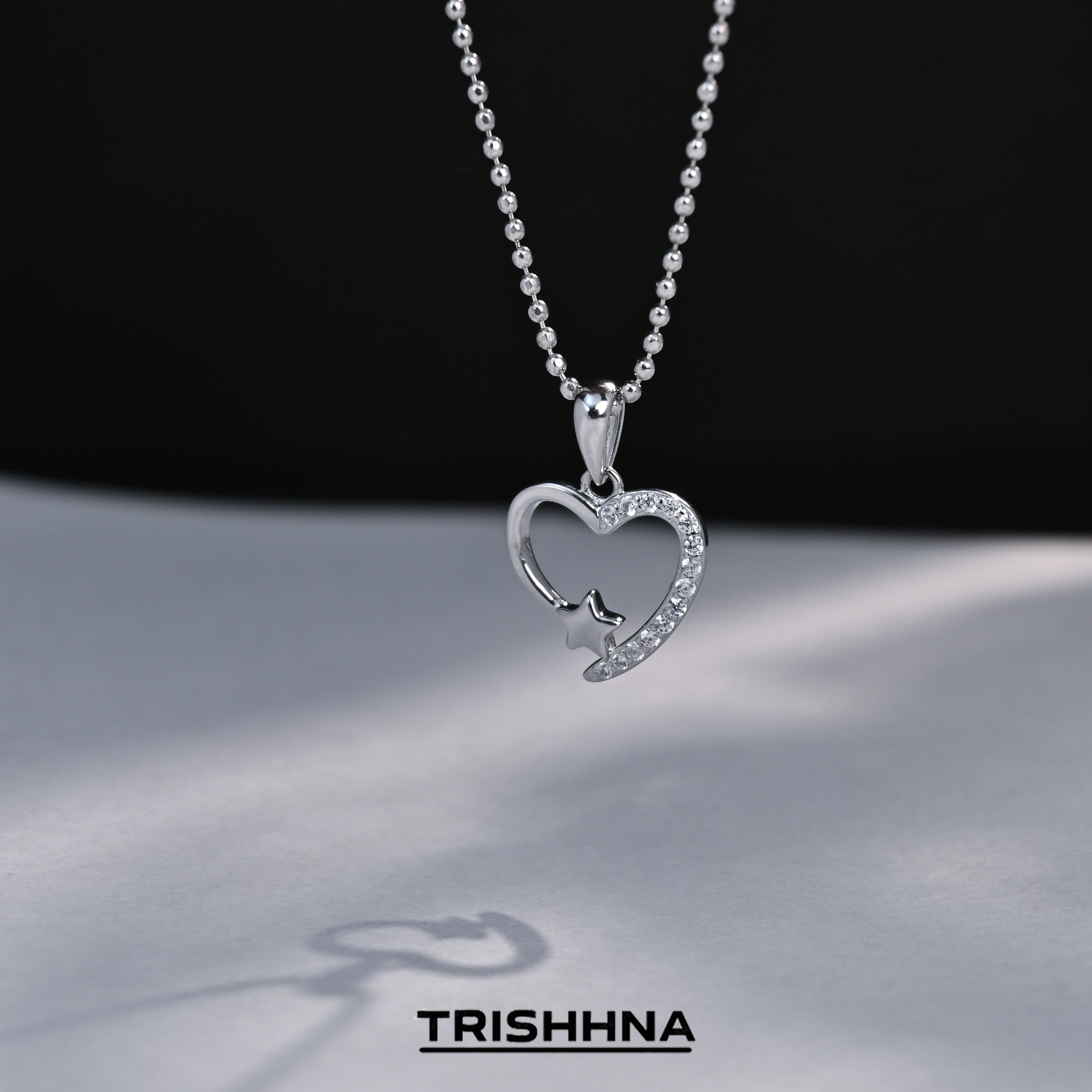 Heart-shaped pendant necklace on a dark background with 'TRISHNHA' branding.