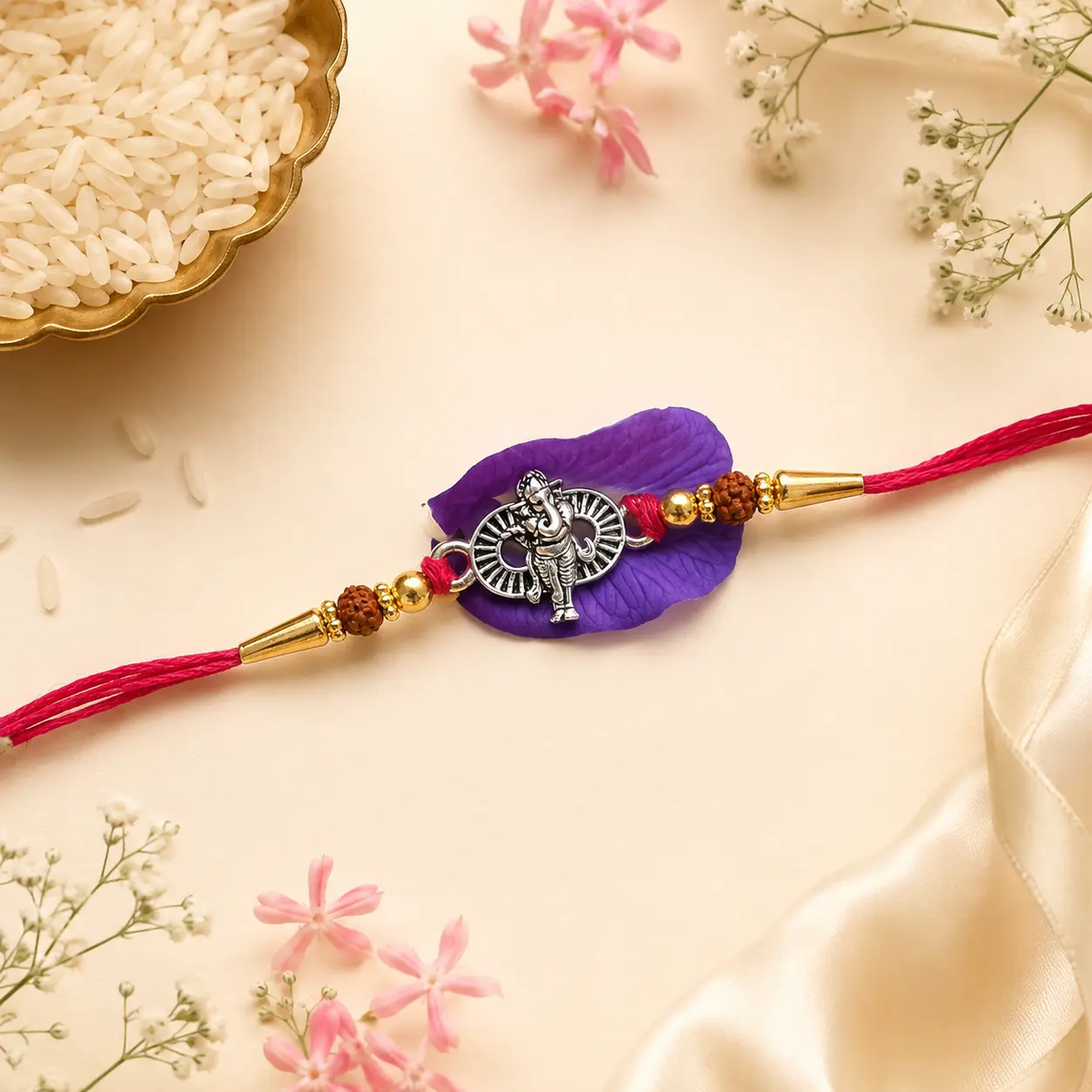 A traditional Indian Rakhi bracelet laid flat on a light cream background, featuring a purple petal, a silver deity charm, red cord, and gold-toned beads. A small gold bowl of white rice and delicate pink flowers are styled around it.