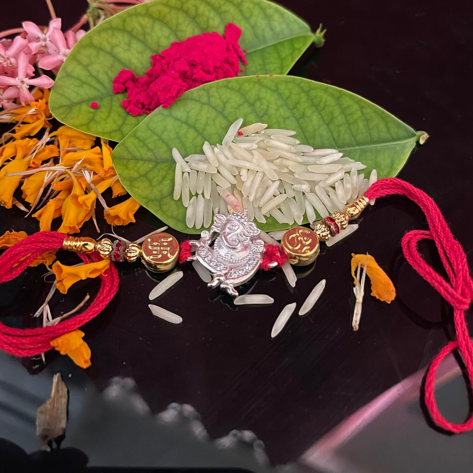 A handmade silver-toned Lakshmi pendant rakhi with red and gold thread details, displayed on two green leaves with white rice and pink and orange flower petals on a dark reflective surface.