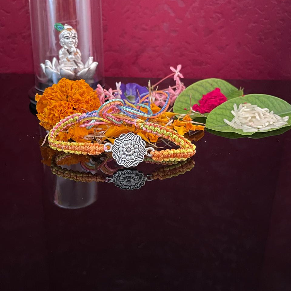 A handmade braided bracelet with a silver mandala charm is displayed on a dark reflective surface, surrounded by marigold flowers, a silver figurine under a glass dome, and green leaves with white flowers against a textured magenta background.