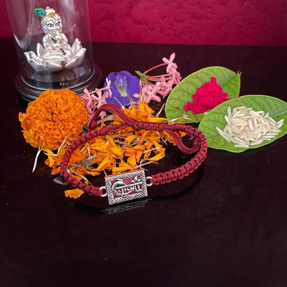 A braided red and gold Rakhi bracelet with a silver 'Swastika' charm, displayed on a dark reflective surface with marigold flowers, a glass dome containing a silver idol, and decorative leaves against a textured dark red background.