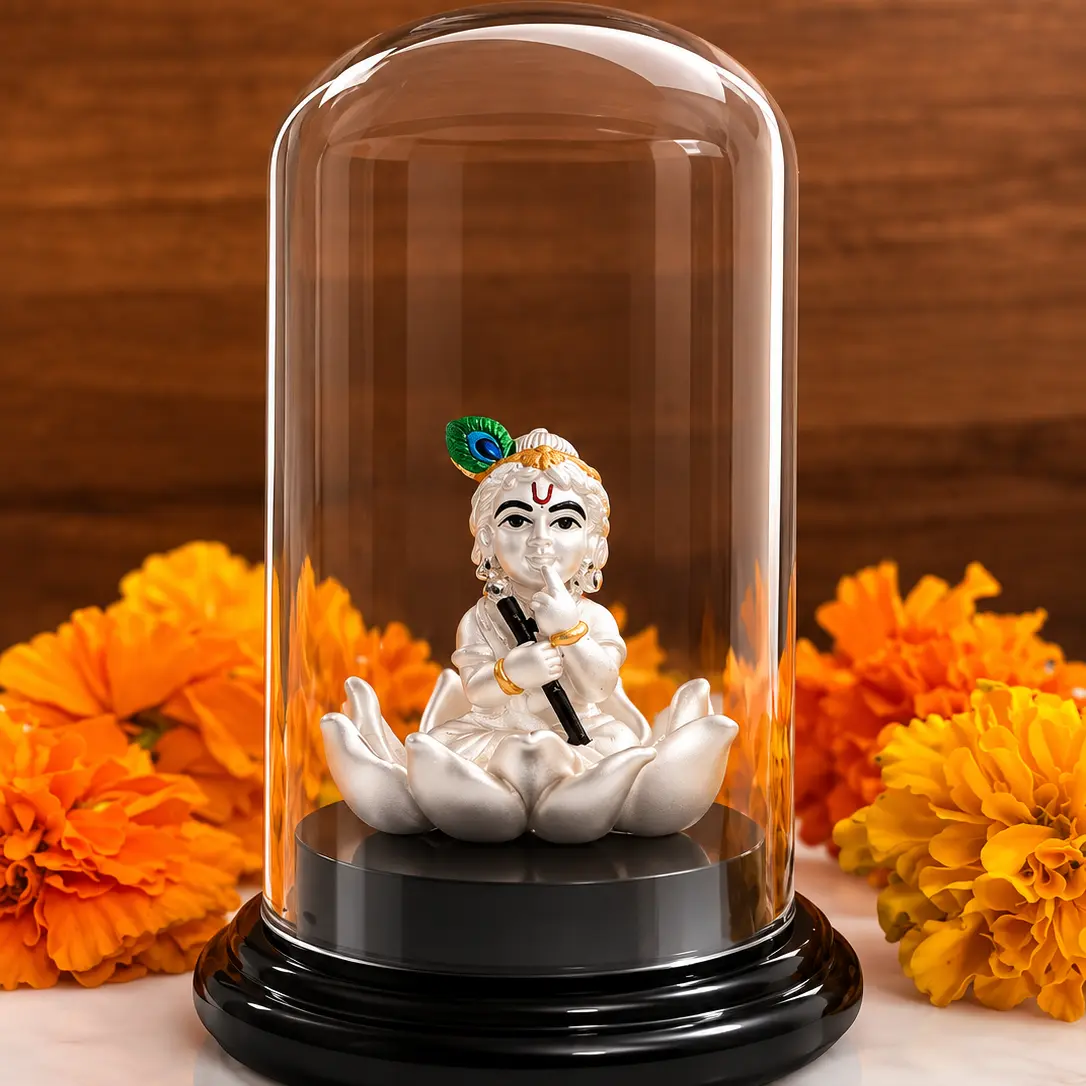 A white Lord Krishna figurine seated in a lotus position under a clear glass dome on a black base, styled with orange marigold flowers on a white marble surface against a wooden background.