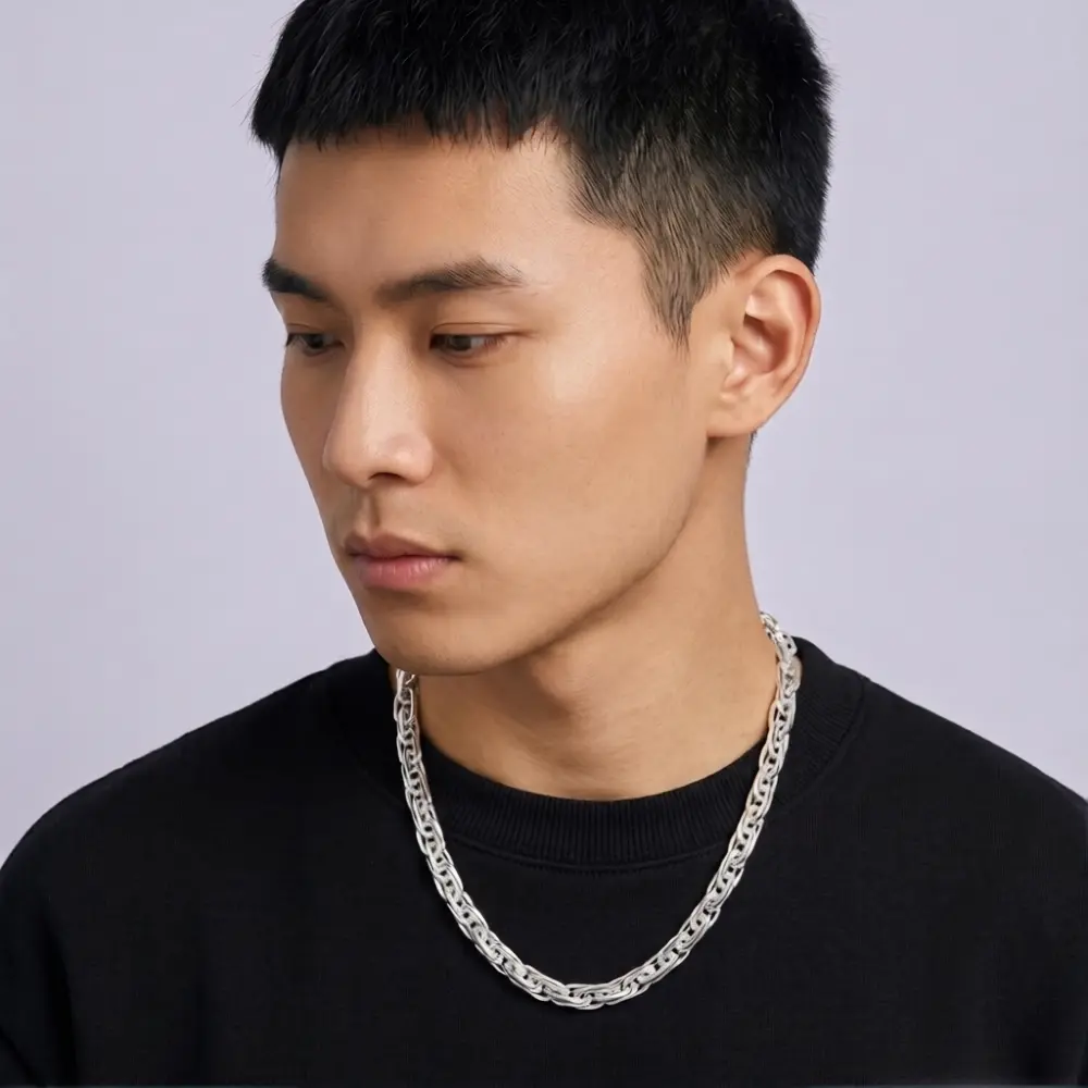 A man wearing a thick silver chain necklace and a black crewneck sweatshirt, shown in a close-up profile shot against a light gray background.