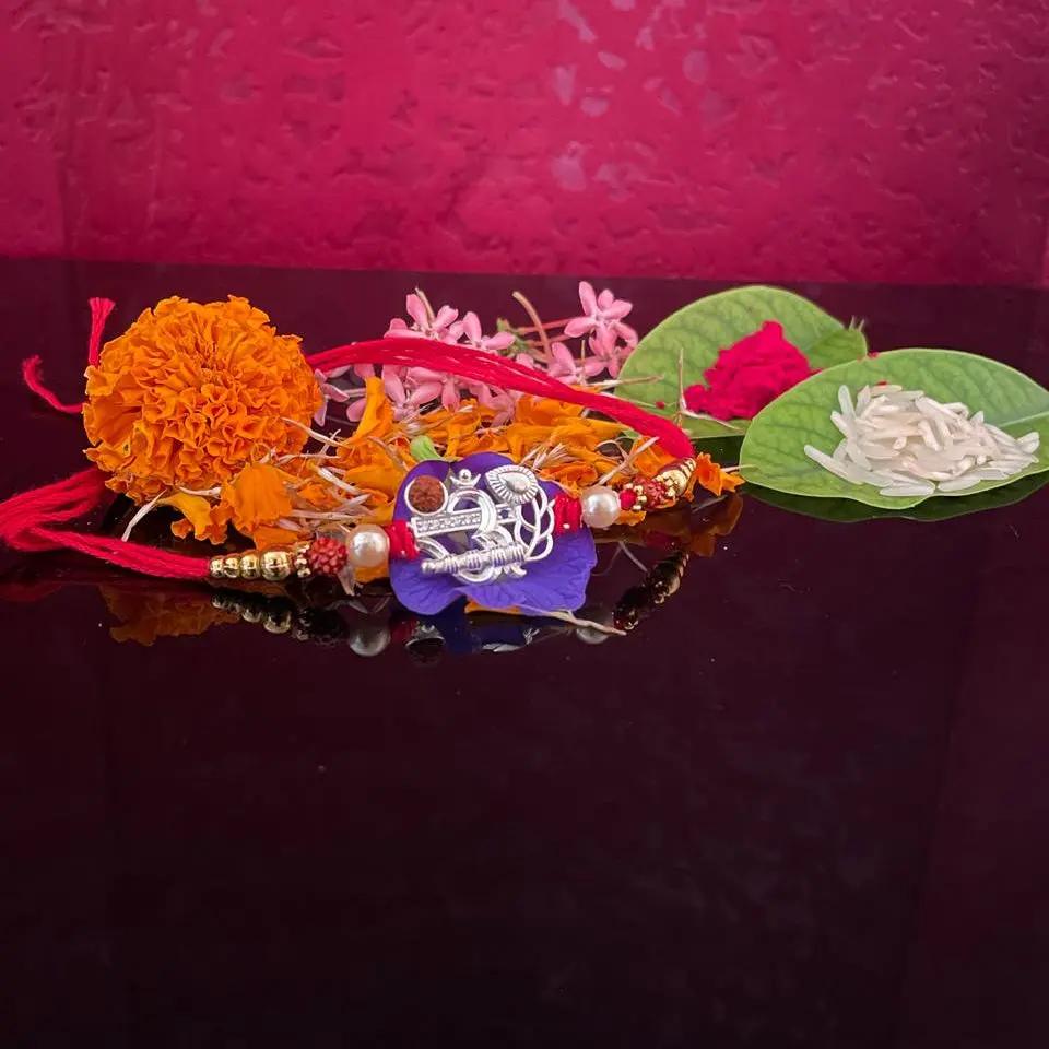 A collection of traditional Indian Rakhi wristlets displayed on a dark reflective surface against a textured magenta-pink background. The Rakhis feature vibrant orange marigold flowers, pink blossoms, green leaves with white rice, red threads, and a central purple Rakhi with a silver foil 'R' charm.