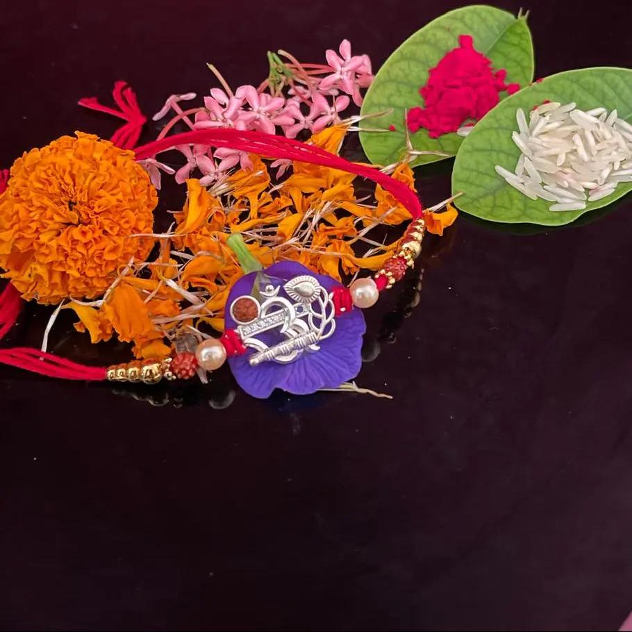 A decorative Rakhi laid out on a dark reflective surface with a textured magenta background. The Rakhi includes a purple lotus charm with silver Devanagari script, pink and orange flower petals, white beads, and two green leaves containing red and white rice.