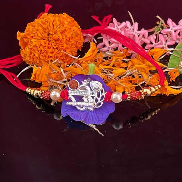 A decorative Rakhi with orange marigold, pink flowers, red thread, and a silver-toned pendant, displayed alongside two green leaves containing rice and red powder on a dark reflective surface against a textured magenta background.