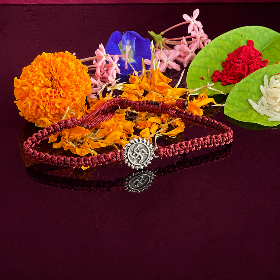 A traditional Indian Rakhi bracelet with a silver-toned circular pendant, displayed on a dark reflective surface alongside marigold flowers, pink blossoms, a blue flower, red powder, and white rice on green leaves, set against a deep burgundy background.
