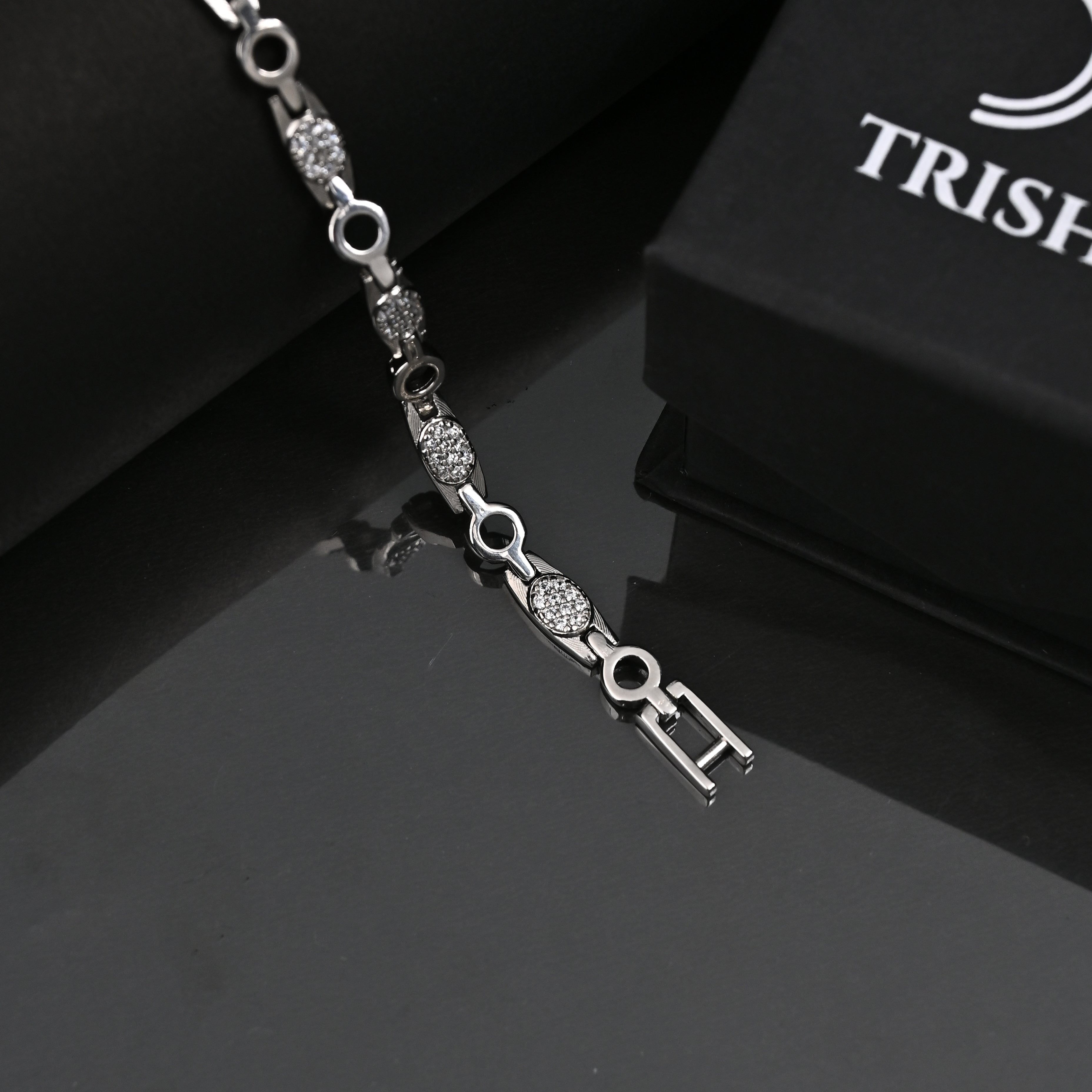 Silver bracelet with key charm on a reflective surface, branded 'Trishhna'.