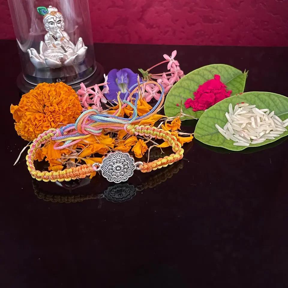 A decorative beaded bracelet with a silver-toned filigree center charm is displayed on a dark reflective surface, surrounded by marigold flowers, pink and purple blossoms, green leaves containing white rice, and a small silver figurine under a clear dome against a deep pink textured background.