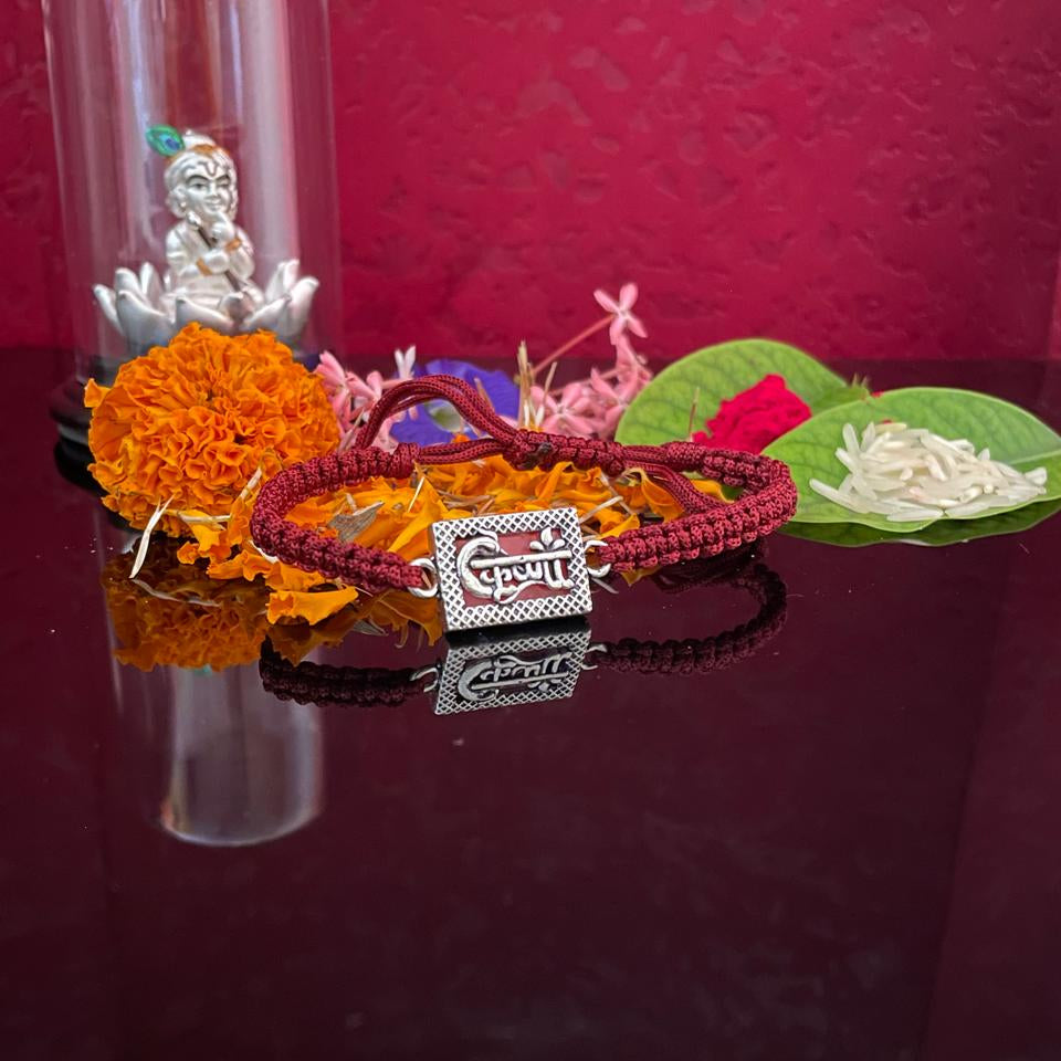 A red braided cord bracelet with a silver-toned rectangular charm engraved with Devanagari script, displayed on a dark reflective surface with orange marigold flowers, green leaves, and a small silver figurine under a glass dome against a deep red background.