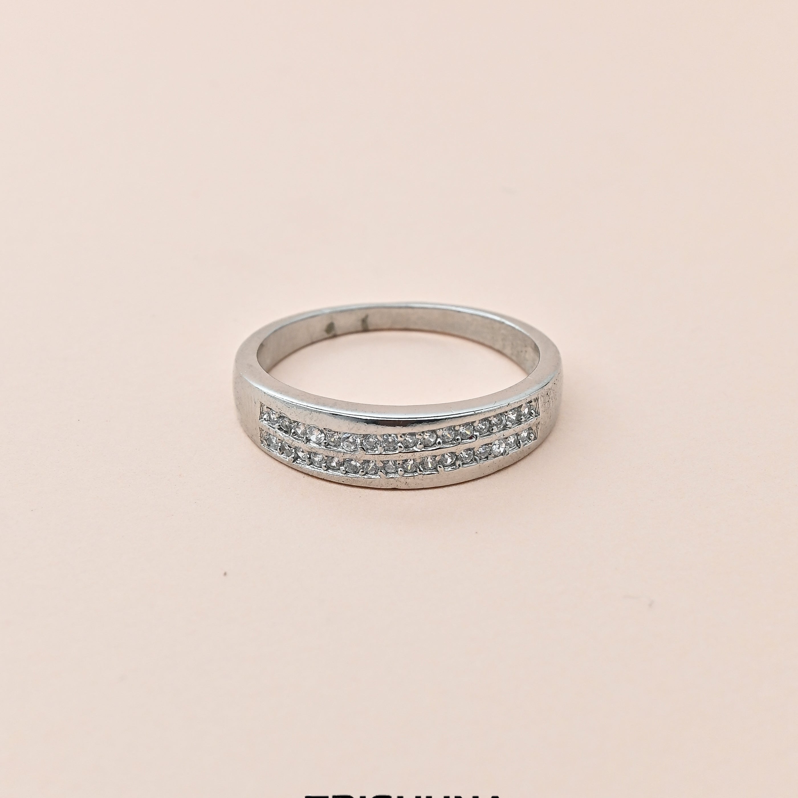 Silver ring with small stones on a beige background, branded 'Trishhna'.