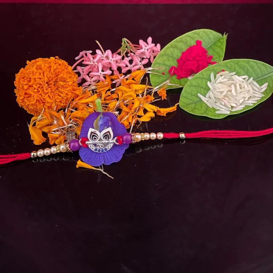 A traditional Indian Rakhi with a purple paper doll charm, decorated with marigold, pink flowers, rice, and a red flower on green leaves, laid out on a dark reflective surface against a textured magenta background.